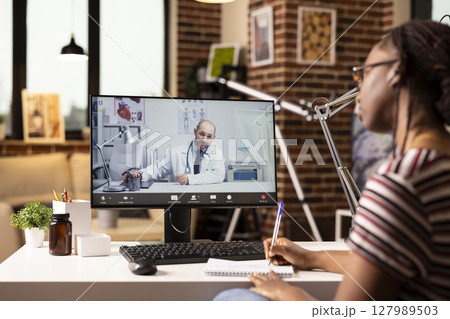Black female patient talking to caucasian doctor on video call, taking notes during remote medical consultation. Woman chatting with male physician, attending virtual health appointment from home. 127989503