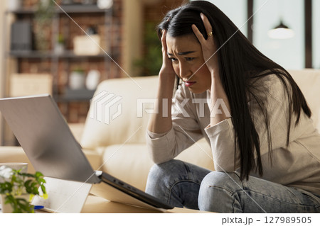 Stressed businesswoman sits on couch, holding head in frustration after reading bad news on laptop. Overwhelmed female entrepreneur feeling tired and emotionally exhausted at home. 127989505