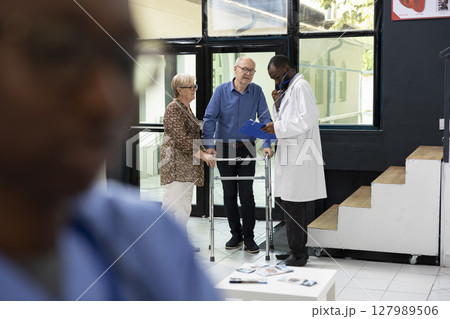 Black medic assisting senior man with reduced mobility in hospital lobby, giving personalized medical care to patient with walking frame. Offering support and advice after thorough checkup. 127989506