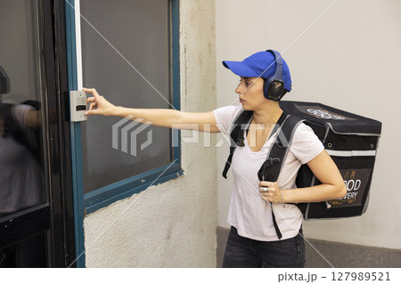 Delivery employee with thermal backpack arrives at the door ringing the bell, approaches a doorway to deliver a takeout meal order. Woman takeaway service courier using the intercom. Delivery employee with thermal backpack arrives at the door ringing the bell, approaches a doorway to deliver a takeout meal order. Woman takeaway service courier using the intercom. 127989521