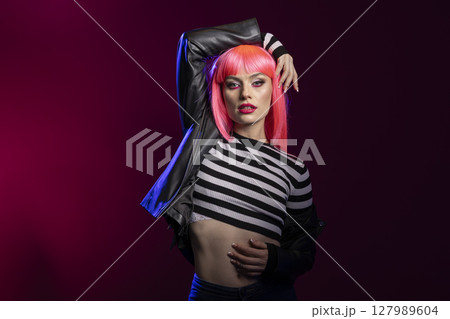 Strong elegant woman projects attitude in studio using bold expression and confident posture. Fierce female model with pink hair, wearing black leather jacket poses against isolated background. 127989604