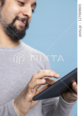 Cheerful and focused young person using his tablet for online tasks, embodying the concept of digital connectivity. Man with long hair browsing the web for modern work or internet communication. 127989605