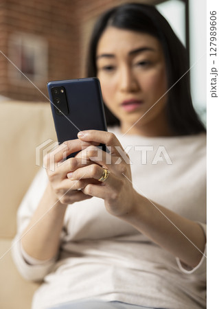 Self employed asian woman relaxing on sofa, using cellphone to chat with vendors, confirming shipment of business products. Remote employee checking social media during work break. 127989606