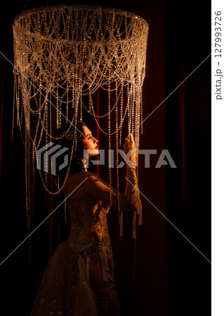 Young woman in vintage corset costume posing under chandelier on dark background Young woman in vintage corset costume posing under chandelier on dark background 127993726