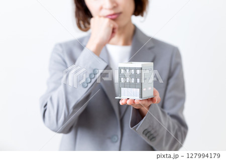 ビルの住宅模型を持って悩むグレーのスーツ姿のミドル女性 ビルの住宅模型を持って悩むグレーのスーツ姿のミドル女性 127994179