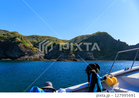 ヒリゾ浜渡船に乗って出港するスキンダイバー、シュノーケラー。 青い海と無人島の大根島、岩礁が見える。 127995691