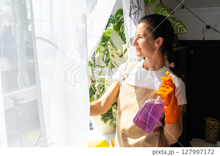 Woman Portrait in rubber gloves and apron manually washes window of house with rag cleaner and mop inside interior with home plants. Restoring order and cleanliness in spring, cleaning servise Woman Portrait in rubber gloves and apron manually washes window of house with rag cleaner and mop inside interior with home plants. Restoring order and cleanliness in spring, cleaning servise 127997172
