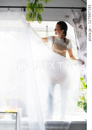 Woman in apron hangs transparent tulle curtains on large attic windows in the house inside the interior with potted plants. Spring cleaning, tidying up 127997191