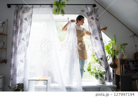 Woman in apron hangs transparent tulle curtains on large attic windows in the house inside the interior with potted plants. Spring cleaning, tidying up 127997194