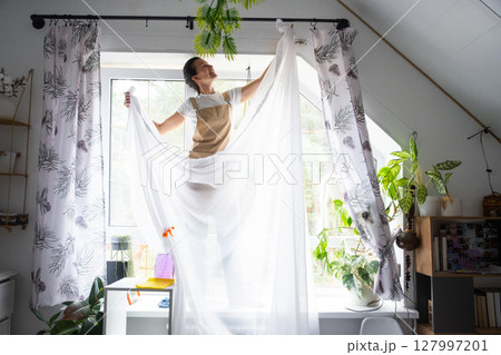 Woman in apron hangs transparent tulle curtains on large attic windows in the house inside the interior with potted plants. Spring cleaning, tidying up Woman in apron hangs transparent tulle curtains on large attic windows in the house inside the interior with potted plants. Spring cleaning, tidying up 127997201