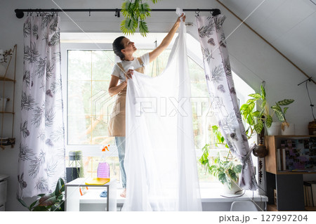 Woman in apron hangs transparent tulle curtains on large attic windows in the house inside the interior with potted plants. Spring cleaning, tidying up 127997204