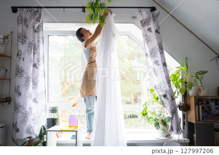 Woman in apron hangs transparent tulle curtains on large attic windows in the house inside the interior with potted plants. Spring cleaning, tidying up 127997206