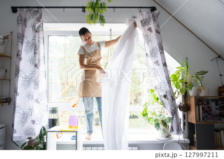 Woman in apron hangs transparent tulle curtains on large attic windows in the house inside the interior with potted plants. Spring cleaning, tidying up Woman in apron hangs transparent tulle curtains on large attic windows in the house inside the interior with potted plants. Spring cleaning, tidying up 127997211