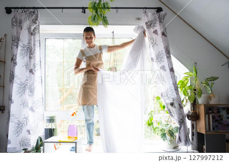 Woman in apron hangs transparent tulle curtains on large attic windows in the house inside the interior with potted plants. Spring cleaning, tidying up Woman in apron hangs transparent tulle curtains on large attic windows in the house inside the interior with potted plants. Spring cleaning, tidying up 127997212