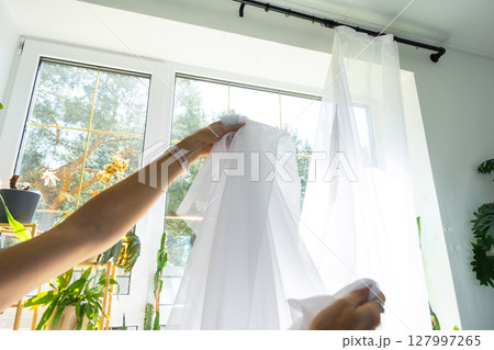 Woman in apron hangs transparent tulle curtains on large windows in the house inside the interior with potted plants. Spring cleaning, tidying up Woman in apron hangs transparent tulle curtains on large windows in the house inside the interior with potted plants. Spring cleaning, tidying up 127997265