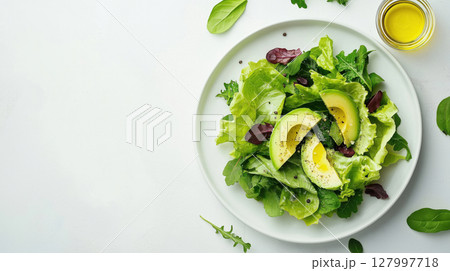 Fresh green salad with sliced avocado, mixed leafy greens, black pepper and olive oil on white plate over light background. Healthy clean eating. 127997718
