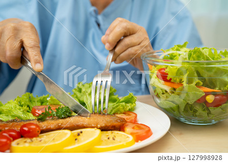 Asian elderly woman patient eating salmon stake and vegetable salad for healthy food in hospital. Asian elderly woman patient eating salmon stake and vegetable salad for healthy food in hospital. 127998928