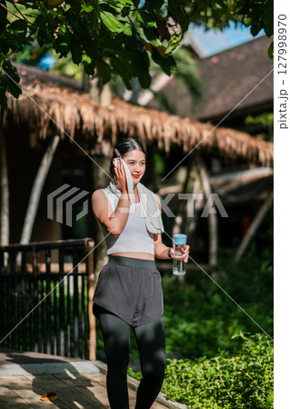 Woman in athletic wear with water bottle, enjoying a sunny day in a green park setting. 127998970