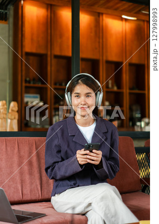 Smiling young woman wearing headphones, holding a smartphone, seated in a stylish cafe with a warm ambiance. 127998993