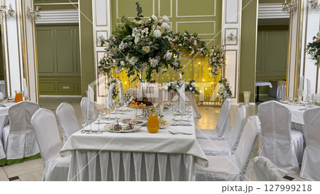 Elegant banquet table with floral centerpiece, fresh fruit, and drinks in luxurious banquet hall. White chair covers, glasses, and fine dining set the scene for a formal celebration or wedding Elegant banquet table with floral centerpiece, fresh fruit, and drinks in luxurious banquet hall. White chair covers, glasses, and fine dining set the scene for a formal celebration or wedding 127999218