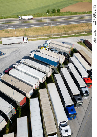 Aerial view of a large truck parking lot at a logistics hub, with multiple commercial semi trucks parked in organized rows. Aerial view of a large truck parking lot at a logistics hub, with multiple commercial semi trucks parked in organized rows. 127999245