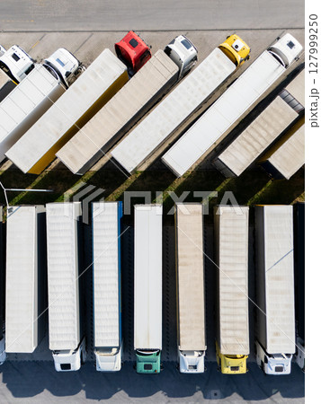 Aerial view of a large truck parking lot at a logistics hub, with multiple commercial semi trucks parked in organized rows. Aerial view of a large truck parking lot at a logistics hub, with multiple commercial semi trucks parked in organized rows. 127999250