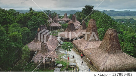 Aerial view of unique custom houses with high, thatched roofs forming a captivating ancient village in a remote area of Sumba island, Indonesia, with local people walking on a path Aerial view of unique custom houses with high, thatched roofs forming a captivating ancient village in a remote area of Sumba island, Indonesia, with local people walking on a path 127999492