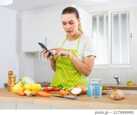 Young housewife using phone to find recipe while cooking dinner Young housewife using phone to find recipe while cooking dinner 127999998
