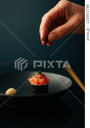 Elegant hand sprinkles dried seaweed over fresh salmon gunkan sushi on a black plate, japanese  128001599