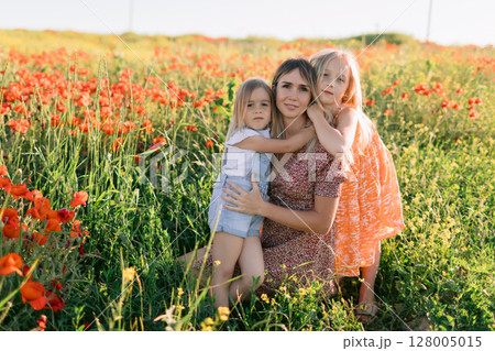 Happy mother with two daughters enjoying picnic in blooming poppy field. Family love concept. 128005015