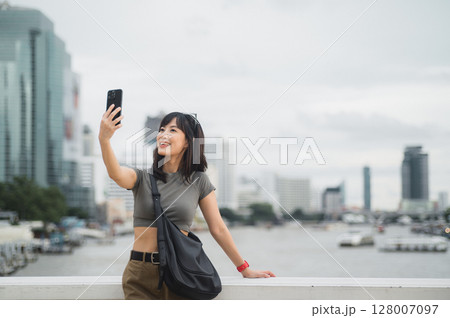 Beautiful young southeast asian woman blogger in 30s taking selfie with smartphone. World travel explorer local cultural experience summer tourism concept at Chao Phraya River Bangkok Asia 128007097