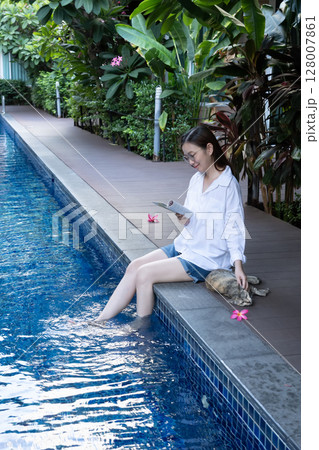 Asian woman reading book relaxing by the poolside, dipping feet in the turquoise water, use hand stroking cat for play with domestic animal on vacation day. Animal therapy, Life with Pets concept Asian woman reading book relaxing by the poolside, dipping feet in the turquoise water, use hand stroking cat for play with domestic animal on vacation day. Animal therapy, Life with Pets concept 128007861