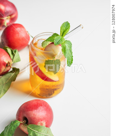 Ice tea with peach, mint in a glass on a white table near window with fresh fruits and sunlight. 128007974