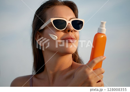 A beauty woman applying sunscreen on healthy skin. Portrait of a female holding a bottle of sun cream and using cream. Skincare product, suntan lotion, or moisturizing solar A beauty woman applying sunscreen on healthy skin. Portrait of a female holding a bottle of sun cream and using cream. Skincare product, suntan lotion, or moisturizing solar 128008355