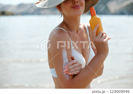 Woman with radiant skin is applying sun cream on her arm, enjoying a sunny beach day, highlighting the importance of skin care and protection. Skin Care Concept. 128009046