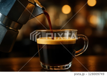 Rich espresso coffee with crema pouring from moka pot into clear glass cup 128009749