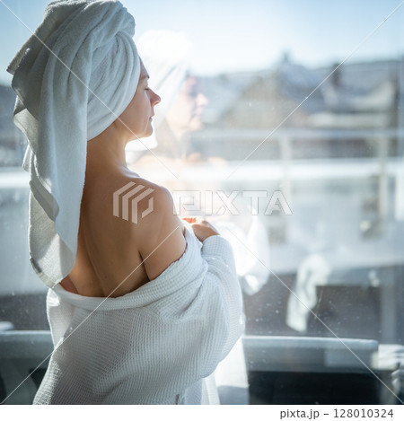 Back view of young woman with towel on her head looking out the window 128010324