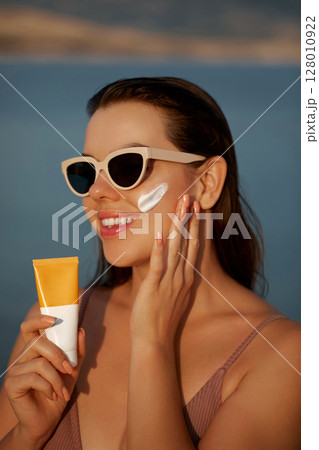 Smiling woman wearing stylish sunglasses applies sunscreen to her face while sunbathing near a serene beach. Promotes skincare, sun protection, leisure, and outdoor wellness Smiling woman wearing stylish sunglasses applies sunscreen to her face while sunbathing near a serene beach. Promotes skincare, sun protection, leisure, and outdoor wellness 128010922