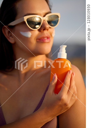 A beauty woman applying sunscreen on healthy skin. Portrait of a female holding a bottle of sun cream and using cream. Skincare product, suntan lotion, or moisturizing solar. A beauty woman applying sunscreen on healthy skin. Portrait of a female holding a bottle of sun cream and using cream. Skincare product, suntan lotion, or moisturizing solar. 128010930