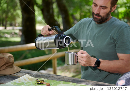 Man Pouring a Drink From a Thermos in a Wooden Outdoor Setting 128012747