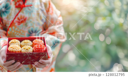 Close up of child in traditional chinese kimono holding festive red box with colorful mooncakes. Banner for mid autumn festival, celebration and cultural traditions. Copy space for text 128012795