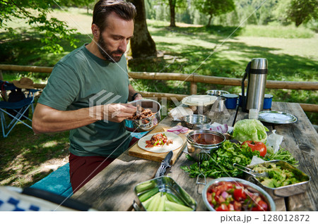 Man Preparing Outdoor Tacos Amidst Nature With Fresh Ingredients on Wooden Table 128012872