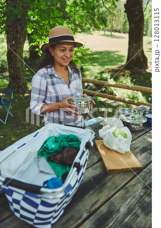 Smiling Woman Outdoors Preparing for Picnic at Wooden Table in Scenic Forest Smiling Woman Outdoors Preparing for Picnic at Wooden Table in Scenic Forest 128013115
