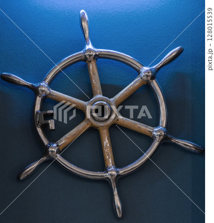 A metallic ship wheel mounted on a blue wall, symbolizing maritime themes A metallic ship wheel mounted on a blue wall, symbolizing maritime themes 128015539