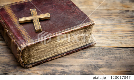 A weathered book rests on a wooden surface, adorned with a golden cross as a symbol of faith 128017847
