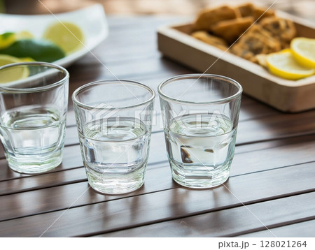 Three vodka shots on wooden table with a plate of snacks in the background, cozy and inviting scene. 128021264