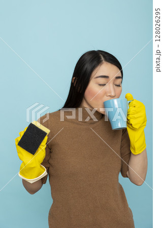 Asian housemaid peacefully sipping coffee while holding sponge, taking relaxing break during household chores. Female janitor with yellow rubber gloves, enjoying calm moment during spring cleaning. Asian housemaid peacefully sipping coffee while holding sponge, taking relaxing break during household chores. Female janitor with yellow rubber gloves, enjoying calm moment during spring cleaning. 128026295