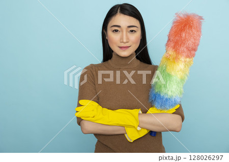 Portrait of confident female janitor wearing yellow rubber gloves and holding feather duster, ready for cleaning duties. Asian housekeeper standing with crossed arms against blue background. Portrait of confident female janitor wearing yellow rubber gloves and holding feather duster, ready for cleaning duties. Asian housekeeper standing with crossed arms against blue background. 128026297