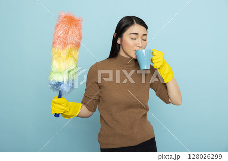 Asian housekeeper holding colorful duster and drinks coffee while taking a break during spring cleaning. Female cleaner standing against blue background enjoying moment of rest from chores. Asian housekeeper holding colorful duster and drinks coffee while taking a break during spring cleaning. Female cleaner standing against blue background enjoying moment of rest from chores. 128026299