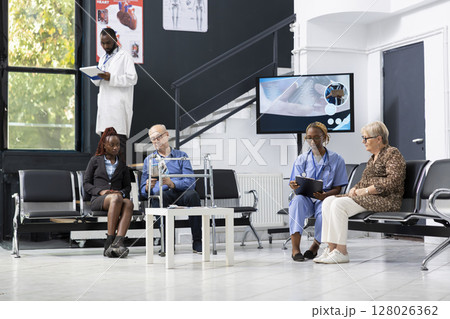 Black healthcare provider discussing with her patients in the hospital lobby, consulting and sharing advice in the waiting room at medical facility. African american nurse chatting with old woman. Black healthcare provider discussing with her patients in the hospital lobby, consulting and sharing advice in the waiting room at medical facility. African american nurse chatting with old woman. 128026362